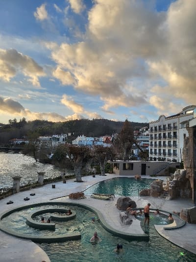 The image captures an outdoor scene at what appears to be a resort or hotel complex, oriented horizontally. In the foreground and midground, a series of intricately designed swimming pools with clear, turquoise water are visible. These pools feature organic, curving shapes, some with rocky edges and others with smooth, light-colored paving surrounding them. Several people are present in the pools, engaged in various leisure activities. Some individuals are partially submerged, while others are standing or sitting near the edges. For instance, one person in dark swim trunks is standing upright in a pool, and another person is sitting on a rock within the water. A distinctive spiraling pool section is visible towards the right side of the image, where more people are seen relaxing in the water. The pool area is well-lit, possibly by natural light complemented by some underwater or pathway lights.
Behind the pools, a large, multi-story building with a light-colored facade and numerous windows and balconies dominates the left side of the image, suggesting it is a hotel or apartment complex. Further in the background, a lush green hillside or mountain, densely covered with trees and vegetation, stretches across the horizon. Scattered buildings, likely part of the same complex or a nearby town, can be seen nestled among the trees on the slopes. A body of water or a wide paved area separates the immediate pool complex from the distant buildings on the hill.
The sky, occupying the upper portion of the image, is a vibrant blue interspersed with large, fluffy clouds. The lighting suggests it is either late afternoon or early evening, as some of the clouds exhibit warm, golden and orange hues, indicative of a sun nearing the horizon, while others appear brighter white or grayish. The overall atmosphere conveys relaxation and enjoyment in a scenic, well-maintained environment. No discernible text is visible in the image.