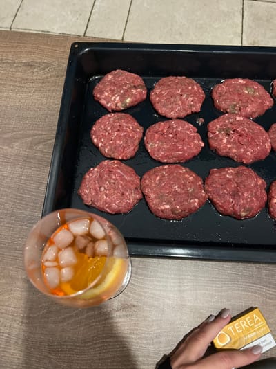A black baking sheet holds twelve raw hamburger patties. Next to it sits a cocktail with ice cubes and an orange slice. A woman's hand, with painted nails, holds a pack of yellow Terea cigarettes. The scene is on a table with a brown surface. The left of the image shows a tiled floor.
