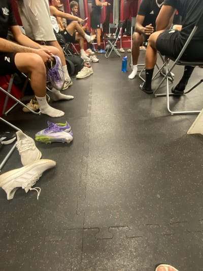 The scene depicts an indoor athletic facility, consistent with a locker room or team meeting area within Lawrence Township, United States. The floor is covered with interlocking black rubber mats featuring small reddish speckles. Dark red or maroon walls are visible in the background, possibly indicating locker bays.
At least six individuals, primarily adult males, are present, with their lower torsos and legs prominently featured. They are seated on black metal folding chairs. Most are wearing athletic shorts (black, white) and socks (white or black); some individuals wear black Adidas slides.
Several objects are scattered on the floor. In the foreground, on the left, a person holds a pair of purple and lime green athletic cleats. An identical pair of cleats lies on the floor directly beneath. Multiple pairs of light-colored athletic sneakers are also on the floor, including a pair of white Adidas sneakers with visible laces. A blue plastic water bottle rests on the floor to the right of the center. A black bag is partially visible under a chair in the mid-ground.
Individuals are generally in a relaxed, seated posture, suggesting a pre- or post-activity gathering. One person in the mid-ground appears to be reviewing papers or notes. There are no overt actions or dynamic interactions taking place; the scene is static, focused on individuals and their personal athletic gear within the facility.