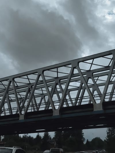 The image captures a close-up, low-angle view of a steel truss bridge under a heavily overcast sky. The bridge's intricate geometric structure, composed of interconnected beams and diagonal supports, dominates the frame. Beneath the bridge, a dark, horizontal surface, likely the road or trackbed, is visible. Further down, a dense line of dark green coniferous trees forms a backdrop, interspersed with glimpses of buildings. In the foreground, the tops of several vehicles, including a silver SUV, are visible, suggesting a road or traffic scenario. The overall impression is one of a cloudy, possibly gloomy day, with the imposing bridge structure as the central element. This scene is likely located in the vicinity of a railway line or road crossing within Sławków, Poland, given the presence of a substantial truss bridge.