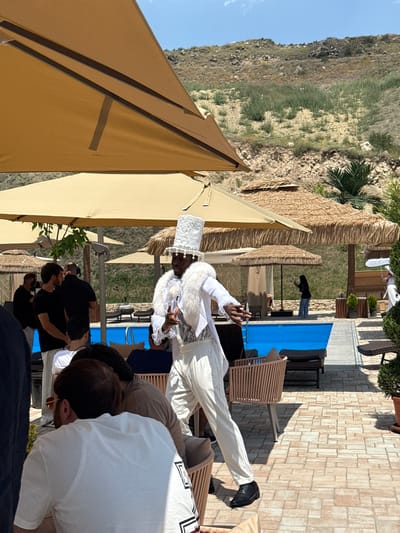 This media file depicts an outdoor scene in Ptghni, Armenia, with a prominent focus on a person dressed in an elaborate white costume, including a tall, ornate white hat, a furry white collar, and a white suit. This individual appears to be in the midst of a performance or celebration, gesturing with their hands and looking towards the camera.
Several other individuals are visible in the background and foreground, some watching the performer, others engaged in conversation. The setting appears to be a resort or a venue with outdoor seating and a pool area. There are thatched-roof structures, lounge chairs, and a blue pool cover. The surrounding landscape shows a hilly, rocky terrain with sparse vegetation under a clear blue sky, suggesting a sunny day.
The overall atmosphere seems festive, possibly a cultural event or a private party. The lighting suggests it is daytime, likely late morning or early afternoon, given the bright sunlight casting distinct shadows. No visible text is present in the media file.