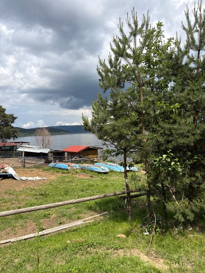 The image depicts a rural lakeside scene in Tsigov chark, Bulgaria, under an overcast sky. The sky occupies the upper portion, filled with a dense cover of varying shades of gray clouds, suggesting diffused light and potentially impending rain. A large body of water, likely a lake, stretches across the mid-ground, reflecting the gray sky and appearing calm. Beyond the lake, a series of gently rolling hills or mountains, partially covered in vegetation, define the distant horizon.
In the mid-ground, along the near shore of the lake, several modest structures are visible. To the left, a structure with a dark roof is partially obscured by trees. Centrally, a cluster of small, single-story shacks or utility buildings feature corrugated metal roofs, some appearing weathered. To their right, a more distinct wooden building with a prominent red gabled roof stands closer to the water. In front of these structures, several light blue, small boats or canoes are resting on the bank, mostly overturned or partially submerged, suggesting they are out of use or stored. A simple, low wooden fence with wire mesh segments delineates this area.
The immediate foreground is characterized by uneven ground, covered with a mixture of green grass and patches of dry, bare soil. Small white wildflowers are scattered throughout the grassy areas. A thick, weathered wooden log acts as a horizontal fence element, traversing the lower mid-section of the frame. To the left of this fence, on a patch of bare ground, a crumpled white tarp or sheet is visible.
Multiple pine trees (conifers) are prominent throughout the frame. A large, mature pine tree is partially visible on the far left. On the right side, several pine trees, along with some deciduous foliage, dominate the foreground and mid-ground, partially obscuring the view of the lake and structures. No people or animals are visible in the scene. The overall impression is one of a quiet, somewhat rustic lakeside environment.