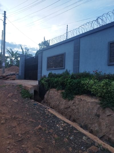 The image shows a dirt road leading to a gated entrance of a property in Othaya, Kenya, located at latitude -0.44 and longitude 36.959. The property is enclosed by a gray wall with a barbed wire fence on top. There is a single window on the wall, and a black gate at the entrance. The road is unpaved and slightly uphill, with a drainage ditch running along the side. There are plants growing along the side of the road and the base of the wall. There are no people or animals present. The sky is overcast. The image is taken from a low angle looking up towards the property entrance.