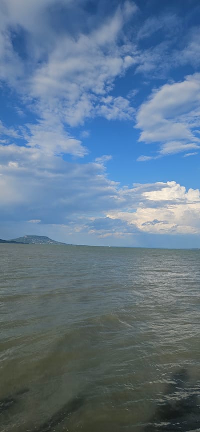 The image depicts a wide expanse of water, likely a lake, with a low-lying landmass on the horizon to the left. The landmass features a prominent hill or mountain with scattered structures and vegetation. The water's surface is rippled, suggesting a gentle breeze. The sky above is a vibrant blue, adorned with numerous white and gray clouds, some of which are large and puffy, while others appear more scattered and wispy. A small white sailboat is visible on the water in the distance. The foreground shows a darker area, possibly the edge of the water with some submerged rocks or debris. The scene appears to be a natural landscape with a tranquil atmosphere. This location is the shore of Lake Balaton in Balatongyörök, Hungary.