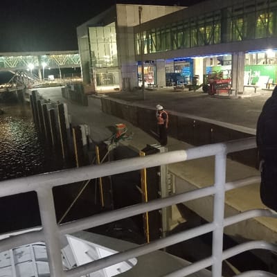 The image depicts a nighttime scene at the ferry terminal in Colonia del Sacramento, Uruguay, viewed from the deck of a vessel. In the immediate foreground, a white metal railing with two horizontal bars and vertical supports is visible, partially obscuring a white structure of the vessel below it.
To the left of the railing, dark water reflects artificial lights, indicating the presence of a pier. Along the water's edge, a paved pier is equipped with several dark, robust vertical bollards or mooring posts. Further down the pier, a single individual stands near the water, wearing a white hard hat, an orange reflective safety vest over dark clothing (possibly a long-sleeved shirt), and dark trousers. They are positioned facing towards the left, slightly away from the observer. Nearby on the pier, some equipment, including a green tarp or cover, is partially visible. Yellow metallic structures, possibly supports or bracing, are seen in and around the water adjacent to the pier.
In the background, a modern, multi-story building with extensive illuminated glass facades stands as the main terminal structure. A glass-enclosed pedestrian skybridge or gangway, a truss-like structure, extends from the terminal building towards the left, connecting to an unseen point over the water. On the paved area beyond the immediate pier, various industrial objects are present, including a red scissor lift or similar construction equipment, and a large green shipping container with partial white lettering, possibly reading "EEN." The entire scene is illuminated by artificial lights against a dark night sky. The upper right corner of the image shows a portion of a person's dark clothing (shoulder and back), indicating the perspective of the photograph.