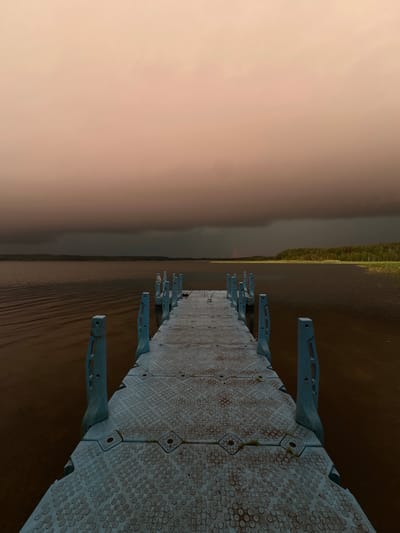 The image depicts a gray, modular plastic dock extending into a calm, brown body of water. The sky above is a muted orange and pink, with dark, heavy clouds forming a band across the horizon. A faint rainbow is visible in the distance, near the treeline on the far shore. The water is rippled, suggesting a gentle breeze. The dock features light blue plastic uprights along its sides. The scene appears to be in a natural setting, possibly a lake or a wide river, in or near Слабодкаўскі сельскі Савет, Belarus. There are no people or animals visible in the image. The overall mood is serene and slightly somber due to the stormy sky.