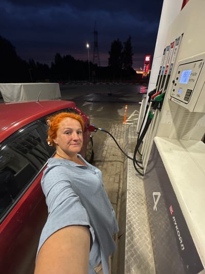 A woman with bright red hair is standing next to a red car at a gas station. She is wearing a light blue patterned shirt. The gas pump display shows "192.9" for price and "3.00" for liters. The Lukoil logo is visible on the side of the gas station. The sky is dark blue, suggesting it is evening or night. There are trees and power lines in the background. The ground is wet, indicating recent rain. The overall scene appears to be a routine stop at a gas station in Mozhga, Russia.