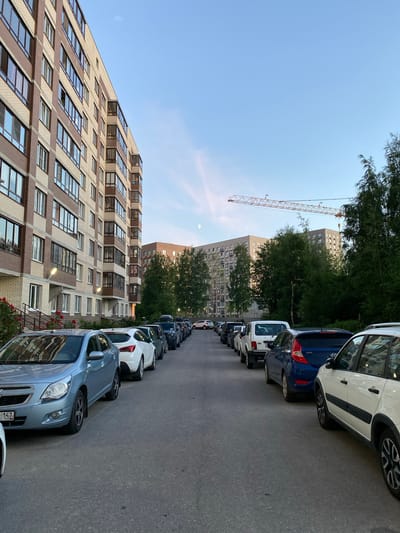 A residential street in Kanisty, Russia, is lined with parked cars and apartment buildings. The buildings, constructed with brick and concrete, appear to be modern and tall, featuring numerous windows and balconies. A construction crane is visible in the distance, suggesting ongoing development in the area. The sky is clear with a hint of pale moonlight visible. The street is paved and shows signs of wear. No individuals are present in the scene. The overall impression is one of a typical urban residential environment.