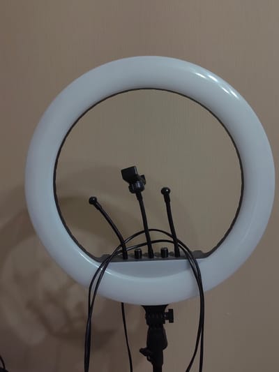 The image shows a ring light with a black phone holder attached, situated against a textured beige wall. Three black flexible arms extend from the base of the ring light; two are positioned lower and shorter, while one is slightly higher and longer, terminating in a small black ball. Another arm, also black and flexible, is mounted centrally and higher up, holding the phone holder. The ring light itself is white and emits a soft, diffused light. Black wires are visible emerging from the base and trailing downwards. The ring light is mounted on a black tripod stand, only the top portion of which is visible. The overall scene suggests a setup for photography or videography. The image does not provide any specific location details within the city of Ostafievo, Russia, or any indication of people, actions, or interactions.
