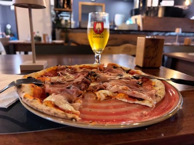 A half-eaten pizza rests on a red-rimmed plate on a wooden table. The pizza is topped with prosciutto, cheese, and what appears to be black olives. A fork lies to the left of the plate, and a knife is positioned to the right, indicating a meal is in progress.
In the background, a glass of Spaten beer is visible, its golden liquid catching the light. The glass bears the Spaten logo. Behind the beer, a wooden table number stand with "13" etched into it is placed. Further in the background, shelves filled with bottles and glassware suggest a bar or restaurant setting. A modern lamp with a white shade stands on the left side of the table, near the pizza. The overall ambiance is casual, likely a dining establishment. The lighting appears to be artificial, indicating an indoor setting, possibly in the evening. The scene conveys a sense of enjoyment and relaxation during a meal.