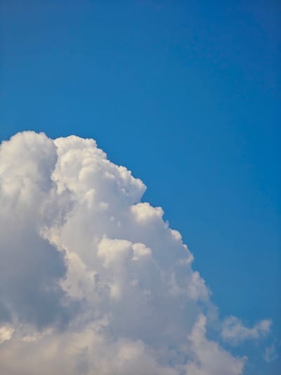 The image shows a partially cloudy blue sky. In the lower left quadrant, a large cumulus cloud dominates the frame, its fluffy white mass contrasting with the deep blue of the sky. The cloud appears bright and textured, with areas of light and shadow suggesting depth. The rest of the image is clear blue sky. No people, objects, or buildings are visible. The scene depicts a natural outdoor environment. The location in Pogodaevo, Russia, is not visually discernible from the image itself.