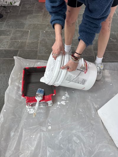 The photograph depicts an individual pouring white paint from a white Benjamin Moore paint bucket into a red paint tray on top of a sheet of clear plastic. The person is wearing a blue sweater and black shorts. Their lower legs and forearms are visible, adorned with tattoos, bracelets, and rings. They are wearing white socks and silver sneakers. A paintbrush is also visible inside the paint tray. The environment includes a gray tile floor, partially covered by the clear plastic sheet. The location is suggested to be Town of Red Hook, United States of America.