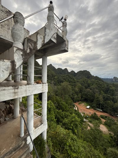 The image shows a concrete viewing platform with a spiral staircase and ornate pillars, overlooking a dense, green forest and hills. The platform appears weathered and aged. A brown dog is lying down on the platform. In the distance, a dirt road winds through the trees, with a few small structures and vehicles visible. The sky is overcast with dramatic clouds. The location is identified as the viewpoint at Wat Tham Khao Phlu, Ban Sadet, Thailand, which is a popular tourist destination known for its impressive limestone karst landscape.