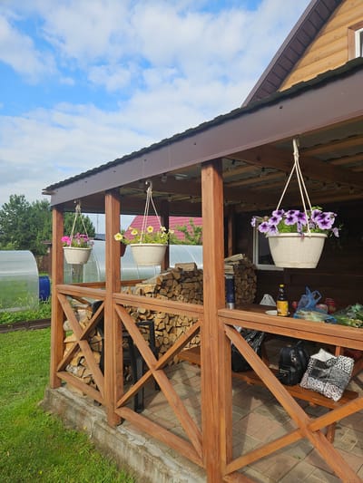 The image shows a wooden gazebo with hanging flower baskets and stacked firewood. The gazebo has a brown roof and is supported by wooden posts and crossbeams. Three hanging baskets with flowers are visible: one with purple petunias, one with yellow flowers, and one with pink flowers. The firewood is stacked neatly on shelves within the gazebo. In the background, a greenhouse and trees are visible. There is also a black handbag and a patterned tote bag on a shelf in the gazebo. The ground is covered with grass and paving stones. The sky is blue with white clouds. The image appears to have been taken outdoors during the daytime, likely in a rural or suburban setting in Gornye Moriny, Russia.