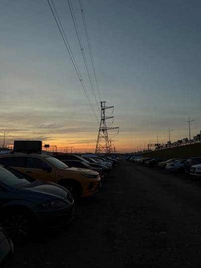 A dimly lit, wide-angle outdoor photograph captures a parking lot filled with cars at dusk. A tall, lattice-like power pylon dominates the right side of the frame, with its wires stretching diagonally across the sky. The sky transitions from a pale blue at the top to warm orange and yellow hues near the horizon, indicating the setting sun. In the foreground, several cars are parked, with their shapes and details obscured by shadows. A yellow taxi with a black sign on its roof is prominently featured on the left side. In the background, more cars are lined up, and a row of industrial-looking buildings or tanks can be seen on a gentle slope. The ground is a dark, gravelly surface. No people are visible in the scene. The location is identified as Severnoye, Russia.