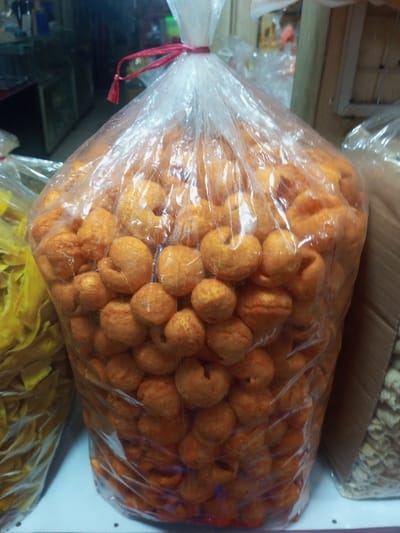 A large, clear plastic bag, tied at the top with a red twist tie, dominates the foreground, filled with numerous small, orange, irregularly shaped puffed snacks. These snacks appear toroidal or spherical with indentations. The bag rests on a white shelf or surface, located within an indoor commercial establishment, likely a market or store in Lê Thanh Nghị Ward, Vietnam. To the left of the main subject, a partial view of another clear plastic bag containing yellow, flat, irregular-shaped food items is visible. To the right, a brown cardboard box or a similar container holding lighter-colored, vaguely defined objects is partially visible. The background is out of focus, suggesting additional shelving or display areas. No individuals, specific actions, or interactions are discernible.