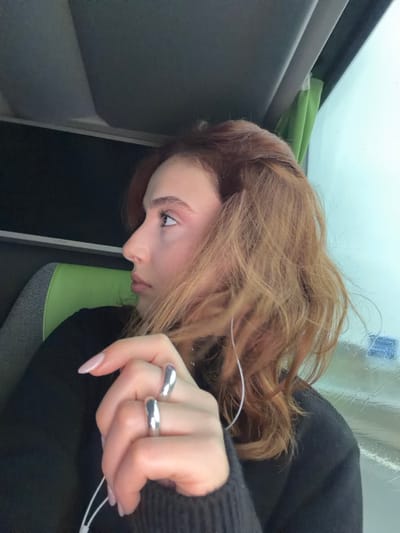The image depicts a young adult female seated inside a vehicle, identified as being in Moineville, France. The subject is shown in profile, facing to the right of the frame, gazing upward and outward through a window. She has wavy, reddish-brown hair with lighter highlights. Her left hand is prominently featured in the foreground, wearing two large, smooth, silver-colored rings on her ring and middle fingers. Her fingernails are manicured with a light, neutral polish. White wired earbuds are visible, with the cord extending from her left ear. She is wearing a dark, possibly black, long-sleeved garment. To her left, part of a green seat or headrest is visible. To her right, a window displays a blurred, indistinct exterior, suggesting motion or an overcast environment, with visible moisture or streaks on the glass. A light green curtain or blind is partially visible at the top right edge of the window frame. The vehicle's interior ceiling, dark grey in color, is visible above her.