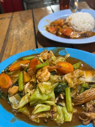 An extreme close-up shot captures two plates of Indonesian food on a wooden table. The plate in the foreground, a vibrant blue, is filled with a stir-fry of vegetables and seafood in a glossy brown sauce. Visible ingredients include large pieces of cabbage, sliced carrots, celery stalks, and several shrimp. To the right, partially visible, is a piece of calamari. In the background, a second plate features a mound of white rice next to a dish with what appears to be chicken and carrots in a similar sauce. The wooden table has a rough, planked surface. The lighting suggests an indoor setting, likely a restaurant. No people are visible in the frame, but a hint of a red object and a blurry glass of amber liquid are present in the background. The scene is located in Boja, Indonesia.