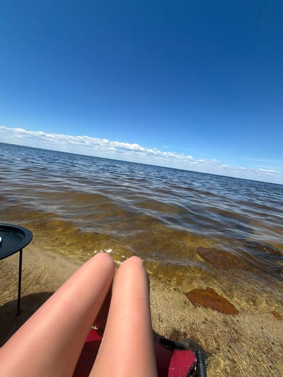 In Lebedivka, Ukraine, on a clear, sunny day, a person's bare legs are positioned in the foreground, extending towards the shallow, clear water of a body of water. The water's surface has gentle ripples, and the sandy bottom is visible, with a few submerged rocks. A small black table with a white object on it is partially visible to the left of the legs. In the background, the water stretches to the horizon, meeting a bright blue sky dotted with a few white clouds. The scene depicts a relaxing moment by the water on a warm day.