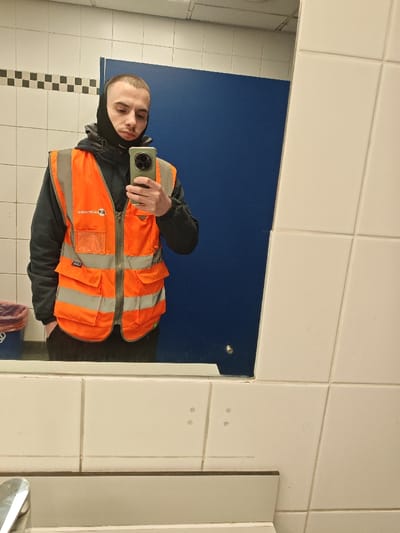 A male individual is depicted taking a mirror selfie in a restroom located in Houghton Regis, United Kingdom. The individual has a short-shaved head and wears a black balaclava covering the lower face and ears, a dark long-sleeved garment, and an orange high-visibility vest with reflective silver stripes and multiple utility pockets, zipped up the front. He holds a mobile phone with a green case, featuring a distinct multi-lens camera array, in his right hand (left in the reflection) to photograph his reflection. The background consists of white tiled walls, a horizontal strip of black and white checkered tiles, and a large blue panel or door. Below the mirror, a white tiled surface with a chrome faucet is visible on the left. A blue waste bin with a red liner is partially visible on the far left, reflected in the mirror.