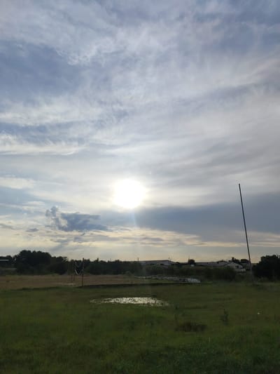This media file depicts a rural landscape in Raiymbek, Kazakhstan, under a cloudy sky with the sun partially obscured. The scene is a flat, grassy field, with a small puddle of water reflecting the sky. In the background, a line of trees forms a dark silhouette against the horizon, and beyond them, several low-lying buildings with white roofs are visible, suggesting a small settlement or farm.
To the left of the frame, a volleyball net is set up, hinting at recreational activity. A tall, slender pole stands vertically in the right side of the image, its purpose unclear. The sun, bright and diffused by the clouds, suggests it is either early morning or late afternoon. The overall atmosphere is serene and natural, with a sense of openness and quietude. No people or animals are directly visible in the frame. There is no visible text in the image.
