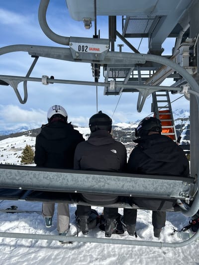 Three individuals are observed from behind, seated on a metallic ski lift chair ascending a snow-covered mountain in Villard-sur-Doron, France. The ski lift structure is visible in the foreground and overhead, marked with the number "002" and the brand "SKIRAIL". A grated metal platform and an orange ladder are part of the overhead lift mechanism.
From left to right:
1. An individual wearing a white ski helmet with "COI" (or similar letters) inscribed on the back, a dark winter jacket, and light grey ski pants. One ski is attached to their right ski boot, which is suspended above the snow.
2. An individual wearing a black ski helmet with goggles pushed up onto it, a small dark camera mounted on the helmet's crown, a dark grey "The North Face" branded winter jacket, and dark ski pants. Their feet are in black ski boots.
3. An individual wearing a black ski helmet with a red and white decal (possibly "BLIZ"), a black winter jacket, and dark ski pants. Their feet are in black ski boots.
All three individuals are facing away from the viewer, looking towards the ascending path. Below them, a vast expanse of white snow covers the ground, with visible ski trails and scattered evergreen trees. In the mid-ground and background, additional snow-covered mountains and valleys extend, with a distant village or settlement visible amidst the terrain. The sky above is partially blue with scattered white clouds, indicating clear weather.