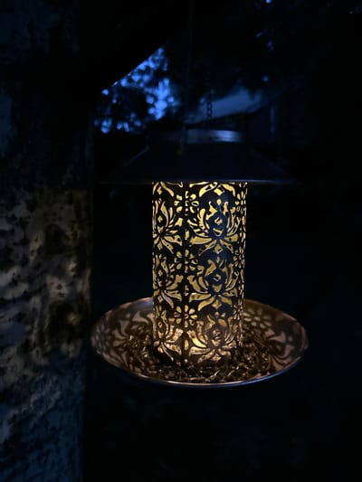A solar-powered bird feeder is hanging outdoors in the dark. The feeder has a black conical roof and a cylindrical body with an intricate, cut-out pattern that illuminates with a warm yellow light. The base of the feeder is a shallow dish filled with birdseed. To the left, a textured, stone-like surface is visible, partially illuminated by the feeder's light. In the background, out-of-focus blue lights suggest distant foliage or possibly a light source. The overall atmosphere is dark and slightly mysterious, with the light from the feeder being the primary illumination. This scene is located in the village of Romashkino, Russia.