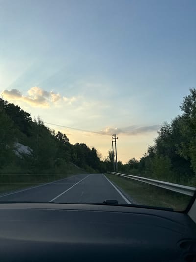 The image captures a view from inside a vehicle, looking out at a road and the surrounding landscape. The scene appears to be a rural or semi-rural setting in Nizhne-Chufichevo, Russia. The road, with white lane markings, curves to the right, bordered by a metal guardrail and lush green trees and vegetation on the left. On the right side of the road, the interior of the vehicle, likely a car door and window frame, is visible, indicating the perspective of a passenger.
The sky is a clear blue with scattered clouds, and the lighting suggests it is either early morning or late afternoon, as the sun's rays are visible casting soft light. The overall mood is peaceful and serene. There is a power line pole with wires visible in the distance, silhouetted against the sky. The foliage is dense and green, with some areas of lighter, almost white, rock or soil visible on a hillside. The image is devoid of any people or text.