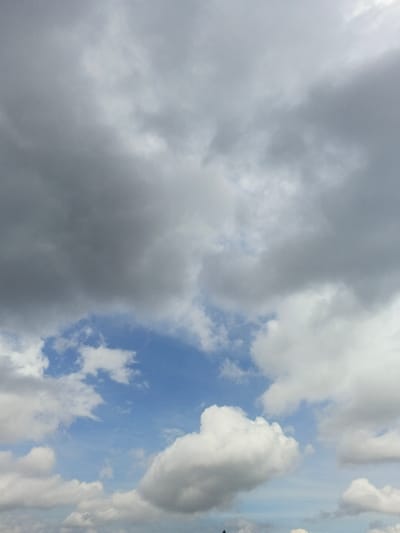 This image presents a view of the sky over Ngọc Tụ, Vietnam. The upper two-thirds of the frame are covered by a dense formation of dark gray and light gray clouds, indicative of overcast conditions. A prominent, irregular opening in the cloud cover in the lower-middle reveals a section of clear blue sky. Beneath this opening, several discrete white cumulus clouds are visible. The bottom edge of the frame shows additional scattered lighter clouds. No terrestrial features, human presence, or man-made objects are discernible.