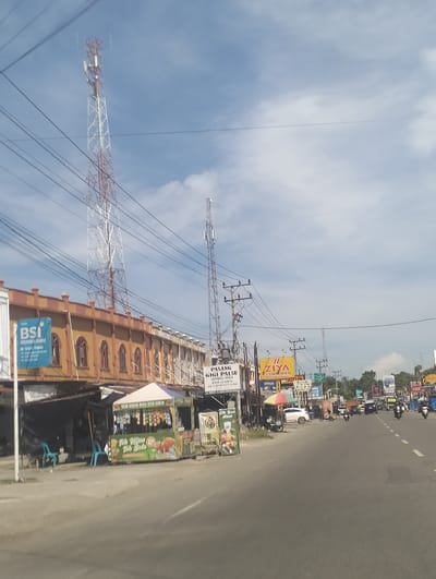 The image depicts a street scene in Grong-Grong, Indonesia, under a partly cloudy sky. A prominent feature is a tall, white and red lattice telecommunications tower located on the left side of the frame. Adjacent to the tower is a long, two-story building with an orange-brown facade, featuring arched windows and a decorative crenellated roofline. A blue sign for "BSI" (Bank Syariah Indonesia) is visible on the left side of the building. In front of the building, a shaded area with a temporary tent structure serves as a small roadside food stall selling beverages and snacks, identified by signage that includes "TEH HIJAU DAN TEH SOLO".

Further down the street, another, smaller telecommunications tower is visible, along with numerous utility poles and overhead wires. A variety of billboards and signs line the right side of the road, advertising different businesses and services, including "ZIYA", "JOTIN", and "PASANG GIGI PALSU". The road itself is paved and appears to be a two-lane street with traffic. Several vehicles are present, including cars, motorcycles, and a truck. People are seen riding motorcycles and walking in the vicinity of the shops. The environment suggests a commercial and urban area with active street life. The overall impression is a typical street view of a bustling town in Indonesia.
