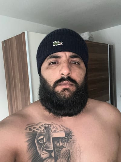 A man with a dark beard and a dark beanie with a Lacoste logo is shown from the chest up. He has a large tattoo on his chest that appears to depict a lion and a cross. He is standing in front of a wooden wardrobe. The scene is indoors. The image appears to be a selfie. The location is Mainburg, Germany.