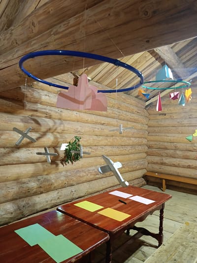 The image depicts the interior of a rustic wooden building, likely a log cabin. The walls and ceiling are constructed from unvarnished, aged logs. Suspended from the ceiling are several decorative mobiles. One mobile features a large blue hoop with pink paper cutouts, while another, smaller mobile, a green hoop, is adorned with colorful geometric paper shapes. Also hanging from the ceiling are several model airplanes, some appearing to be made of wood or painted material. In the foreground, two dark wooden tables are visible. The table on the right has several sheets of paper on it, including yellow and white, along with a dark marker. The table on the left has two sheets of light green paper. A simple wooden bench is positioned against the back wall. The overall atmosphere suggests a room designed for children's activities or a recreational space within a natural or rural setting, specifically in the village of Molochne, Russia. No people are present in the image.