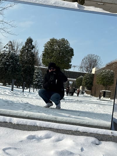 This image captures a reflection in a large, vertical mirror-like surface, situated outdoors in a snow-covered environment within the city of Plodorodny, Russia. The scene is illuminated by bright daylight under a clear blue sky.
In the central foreground of the reflection, a person is depicted in a low squatting stance. This individual wears a black winter jacket, dark blue jeans, white sneakers, a black beanie, and black sunglasses, with gloved hands. They are holding a black smartphone directly in front of their face, positioned to capture a self-portrait of their reflection.
The background, visible behind the primary subject within the reflection, features a park-like setting covered in a layer of fresh snow. To the left, several snow-laden evergreen trees and other bare-branched trees are present, alongside a row of dark, vertical posts protruding from the snow. To the right, a large, dark-textured architectural structure, possibly a wall with a central archway, spans horizontally. Further to the right, a multi-tiered stone wall with some small, rounded green shrubs is visible. Several indistinct figures, appearing to be other pedestrians dressed in winter clothing, are walking along a distant snowy path near the architectural structure.
The reflective surface is bordered by a narrow metallic strip at the top and rests upon a low, snow-covered stone ledge at the bottom. The photograph capturing this reflection is taken from a low vantage point, looking upwards at the reflective surface.