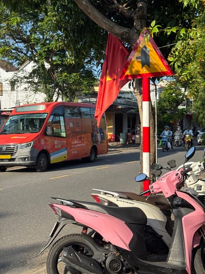 The image depicts a daytime street scene in Hội An Đông Ward, Vietnam. In the immediate foreground, on the right, two Honda Vision scooters are parked; the closest is pink, and the one behind it is white. To their left, a red and white striped pole supports a yellow triangular warning sign indicating a side road merging from the left. A red Vietnamese flag with a yellow star is tied to a tree branch above the pole, hanging downwards.

On the asphalt road, a red minibus, branded with "SHB" and displaying Vietnamese text including "Tiết kiệm hôm nay," travels right-to-left. Its license plate, "50W-276.84," is visible on the front. Ahead of the parked scooters, a line of at least three individuals on motorbikes, wearing helmets and light-colored attire, ride in the same direction. The background features various multi-story buildings, including a prominent white structure on the left and commercial storefronts on the right. Lush green trees line the street, and the scene is illuminated by bright sunlight.