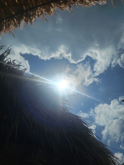 The image shows a view looking upwards towards a bright sky with puffy white clouds. The sun is shining brightly, creating a lens flare effect. In the foreground, parts of a thatched roof are visible on the left and right sides of the frame. The thatch appears to be made of dried palm leaves. The scene appears to be outdoors in Key Largo, United States of America. No people are visible in the frame.