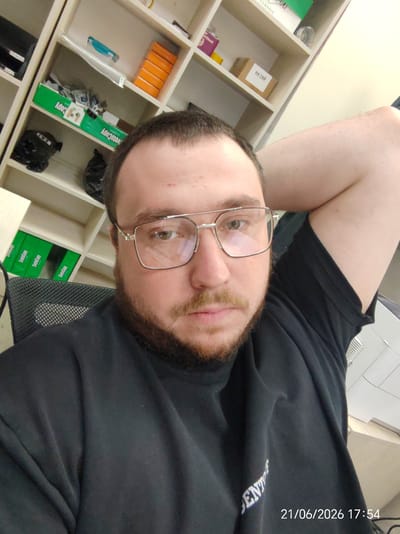A bearded man with glasses is taking a selfie in an office environment. He is wearing a black t-shirt and has his arm resting behind his head. In the background, there are shelves filled with office supplies and boxes. A date and timestamp "21/06/2026 17:54" are visible in the bottom right corner of the image. The location is Prikubanskii, Russia.