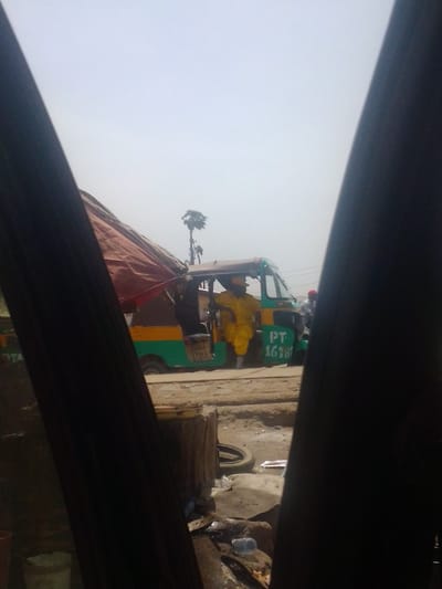 The image captures an outdoor scene in Mista Ali, Nigeria, viewed through a narrow dark opening, possibly a vehicle's window frame. A green and yellow three-wheeled vehicle, identified by "PT 1678" on its side and "PTA" partially visible on the left, is centrally positioned on a dirt road. A male individual, clad in a bright yellow full-body garment resembling a protective suit or uniform, is seated in the driver's section of the vehicle, with his right leg extended out of the open door. His head is covered by a hood. Behind the three-wheeler, two other individuals are partially visible, one wearing a red head covering. The immediate foreground features a partially obscured structure covered by a reddish-brown tarp. The ground consists of loose dirt and mud, strewn with debris including a discarded tire, plastic bottles, and a wooden block. In the background, a tall palm tree rises against a hazy, light grey sky.