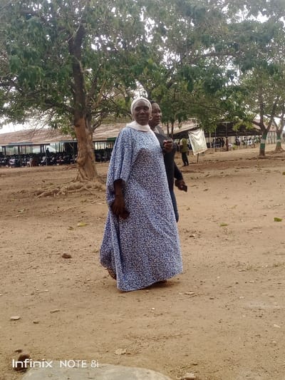 The image displays an outdoor scene in Ayere, Nigeria, characterized by a dry, reddish-brown dirt ground. In the foreground, an adult female with dark skin wears a white head covering and a long, loose-fitting blue dress with a white floral or speckled pattern. She holds a dark object in her right hand and is positioned slightly left of center, looking towards the viewer. Behind and to her right, a second individual, partially obscured, wears dark clothing and appears to be walking in tandem. The mid-ground is dominated by large trees with green foliage and visible trunks. In the background, a long, single-story building with a rusty corrugated metal roof is visible, featuring an open side revealing numerous people seated in rows, suggesting a public gathering or event. A white banner or sheet is visible near a background structure. The overall environment is open, dusty, and contains scattered vegetation and additional distant figures.