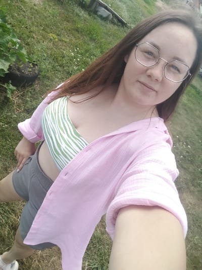 A woman is taking a selfie outdoors. She is wearing a green and white striped bikini top, grey shorts, and a loose-fitting pink shirt that is unbuttoned and worn open. She has long brown hair and is wearing large, geometric-framed glasses. The background appears to be a grassy yard with some foliage to the left. The lighting suggests it is daytime. The overall mood seems casual and relaxed. No text is visible in the image.