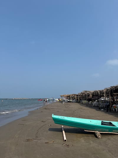 The scene depicts a public beach in Antón Lizardo, Mexico, under a clear blue sky with minimal haze. The foreground features dark grey-brown sand, upon which a teal-colored wooden boat rests, supported by two wooden logs. A black plastic crate is visible inside the boat. To the left of the boat, a light-colored pipe or pole lies on the sand, and a rope extends from the boat towards the ocean.
To the right, a long line of thatched-roof palapas (shelters) extends along the beachfront. Beneath these palapas, numerous white plastic tables and chairs are arranged, with several individuals seated and engaging in dining or relaxation. Further down the beach, multiple smaller wooden boats are pulled onto the sand near the palapas.
The ocean, visible on the left, is a blue-green color with small waves reaching the shore. A rock jetty or breakwater extends into the water on the far left. Several individuals are in the water, swimming or wading. A red inflatable tube, possibly used for water recreation, is visible in the water with people on it. A few individuals are also walking along the wet sand near the water's edge. Birds are visible in the distant sky above the water.