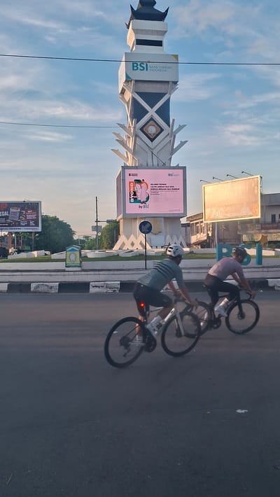 This is a vertical, eye-level, outdoor, daytime shot featuring two cyclists on a paved road in front of a prominent tower and billboards. The tower, likely a landmark, is white and black with decorative elements and displays a large digital billboard at its base. The billboard shows an advertisement for "BYOND by BSI" and congratulates "CALON JEMAAH HAJI 2024" (prospective Hajj pilgrims). To the left of the tower, another billboard advertises "ROKOK ILEGAL" (illegal cigarettes). To the right, a large rectangular billboard is illuminated by the sun. In the background, several buildings are visible. The sky is a pale blue with some cloud cover and is crossed by two dark wires. The road in the foreground is dark asphalt with white and gray markings. The two cyclists are riding side-by-side, with the cyclist on the left wearing a teal jersey and helmet, and the cyclist on the right in a pinkish jersey and helmet. Both are wearing cycling shorts and appear to be in motion. The environment suggests a city setting in Lambaro, Indonesia, possibly a public square or intersection given the tower and multiple billboards.