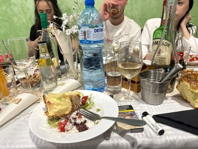 The image shows a cluttered table at what appears to be a social gathering. Several people are partially visible around the table, including a woman with dark hair looking at her phone, a man in a white shirt drinking from a glass, and another woman with dark hair leaning on her hand. Bottles of water and alcohol (including a Jameson whiskey bottle) are prominent. A plate with a piece of food that looks like bread, salad, and cheese sits in the foreground, along with a fork. There's a pack of cigarettes and a lighter on the table, along with various glasses, nuts, and what looks like small party favors. The location appears to be in Rozovo, Bulgaria, based on the "DEVIN" water bottle label and the text on the cigarette packaging. The lighting is bright, and the background is a light green wall.