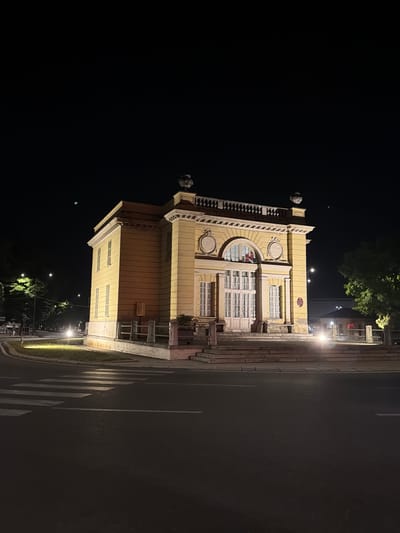 A nighttime shot shows a building with yellow facade, ornate white trim and a red tiled roof. The building has arched windows and a double door in the center with tall glass panels. A set of stone steps leads up to the entrance. The building is located in Parma, Italy, at the intersection of Via Emilia Est and Viale Rustici. A pedestrian crossing is visible in the foreground. The street is illuminated by streetlights and headlights from unseen vehicles. Trees line the street on the left side of the building. The sky is dark and clear.