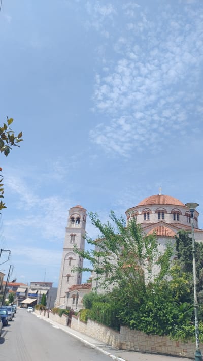 The image depicts a street scene in Νέα Μηχανιώνα, Greece, dominated by a large, stone church with red-tiled roofs and a prominent bell tower. The church features arched windows and crosses atop its domes. A street lined with parked cars and buildings runs alongside a stone wall in front of the church. Lush green trees and foliage are visible in the foreground and around the church. The sky above is a clear blue with scattered white clouds.