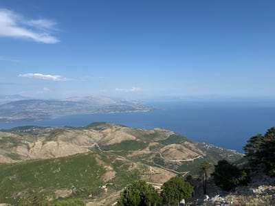 The image captures a panoramic view from a high vantage point in the Kassopaia Municipal Unit, Greece, overlooking a vast expanse of the Ionian Sea and the surrounding mountainous terrain. The foreground features rugged, dry hillsides dotted with sparse vegetation and a few trees. Winding roads are visible, snaking their way up and across the slopes.

In the midground, the landscape transitions to greener, more densely vegetated hills, still characterized by their undulating contours. A body of water, likely a bay or inlet, is visible further back, separating the immediate mountainous region from a distant coastline.

The background is dominated by the deep blue of the Ionian Sea, stretching out to the horizon. Across the water, a long, irregular coastline with mountainous features is discernible, appearing hazy in the distance. The sky above is a clear, bright blue, with a few wispy white clouds scattered across the upper portion of the frame. The scene conveys a sense of natural beauty and remote, rugged topography.