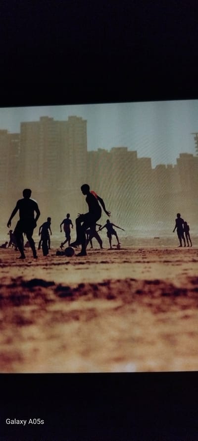 The image displays a vibrant scene of several individuals, likely children or young adults, playing football (soccer) on what appears to be a sandy or earthy ground, possibly a beach or a dusty field. All figures are rendered as dark silhouettes against a bright, hazy sky, suggesting either sunrise or sunset. Their dynamic poses – running, kicking, and jumping – convey energetic movement and engagement in the game. A football is visible near the feet of some players.

In the mid-ground, a body of water is visible, merging into the distant horizon with the hazy sky. Further in the background, a distinctive silhouette of tall buildings or a city skyline can be seen against the soft, diffused light of the sky. The light source is behind the subjects, creating stark, dramatic outlines of the figures. One player has a faint hint of red visible on their clothing. The overall mood is one of active recreation and youthful exuberance.

On the far right edge of the original image (which appears to be a photograph of a screen), the text "Galaxy A05s" is visible in white, indicating the device that captured the image being displayed. While the provided context is Zavoy, Bulgaria, the image itself shows a scene that could be typical of many coastal or lakeside areas with a distant urban setting, without specific identifying landmarks for that location.