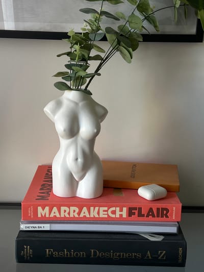This image depicts a still life arrangement located in La Garenne-Colombes, France. A white, unglazed ceramic vase, sculpted as a headless, armless female torso from the waist up, stands centrally. Several green eucalyptus branches protrude from the vase's opening at the neck. The vase rests on a stack of four books. The top book has a coral-red cover with "MARRAKECH FLAIR" and "Marina Berenson" and "Assouline" printed in white text. A tan-brown book with "LOUIS VUITTON" visible on its spine is partially visible behind the vase, also resting on the coral-red book. Beneath the coral-red book is a book with a white spine displaying "DIEYNA BA 1". The bottom book has a black cover with "Fashion Designers A-Z" and "The Collection of the Museum at FIT" and "Taschen" printed in gold text. To the right of the vase, positioned on the coral-red book, is a white Apple AirPods charging case. The entire arrangement is situated on a reflective, clear surface, likely glass. The background consists of a plain, light beige wall, with a thin black frame visible horizontally along the top edge of the image, suggesting a framed object or mirror. No people are present, and no active interactions are depicted.