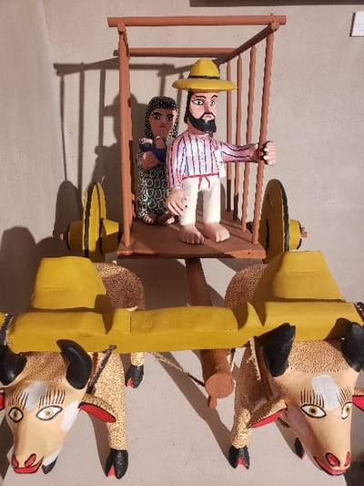 A colorful folk art sculpture depicts a whimsical scene. Three painted bovine figures with black horns pull a yellow-framed cart. Inside the cart, a man with a yellow hat hangs upside down, holding onto a horizontal bar. Beneath him, a woman sits in a patterned dress. The entire piece is brightly colored and appears to be made of wood.