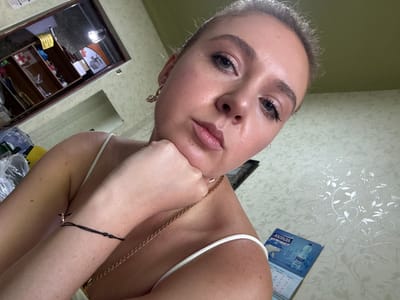 A close-up, slightly upward-angled selfie features a young Caucasian woman with blonde hair pulled back and light makeup. She is wearing a white strap top and gold jewelry, including a pendant necklace and a hoop earring with a stone. Her left hand rests on her chin, and her right arm is bent, with a black string bracelet visible on her wrist. The background shows a shelf with various items, including a red cross symbol, a fan, and some bottles, along with a patterned wallpaper. A calendar with an image of a polar bear is visible in the lower right corner. The lighting suggests an indoor setting.