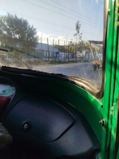 The image presents a street view in Keekonyokie ward, Kenya, captured from the interior perspective of a green-bodied vehicle, likely a tuk-tuk. The foreground displays the vehicle's dark gray dashboard with a visible keyhole or ignition switch on the left, and the lower portion of a dusty windshield fitted with two horizontal wiper blades.
The scene outside features a paved road extending into the distance, curving slightly to the right. Along the left side of the road, a line of irregularly spaced wooden posts forms a fence. Behind this fence, several single-story buildings with light-colored corrugated metal roofs and walls, some appearing green or blue, are visible, interspersed with dense green foliage. Overhead, multiple power lines are discernible against a bright, clear sky with scattered white clouds. Further down the road, on the right, two distinct yellow tuk-tuks are observed; one is closer and partially obscured by atmospheric haze, while the other is further in the distance. A tall, slender tree stands on the right side of the road in the background. No people are explicitly visible. The scene is illuminated by natural daylight.